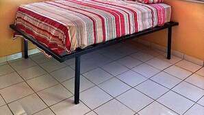 Una cama individual con colcha a rayas en una habitación con paredes anaranjadas, ventilador de techo y ventanas con persianas.