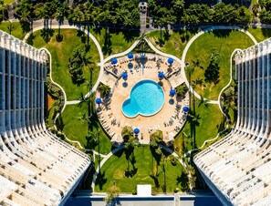 Luftaufnahme eines Hotels mit einem zentralen Swimmingpool, umgeben von Liegebereichen und Palmen.