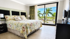 A hotel room with a bed, a desk, a TV, and a balcony with a view of palm trees and the sea. - Luxury Beach Resort Marco Island Condo | Ocean Views, Pool & Beach (Marco Island)