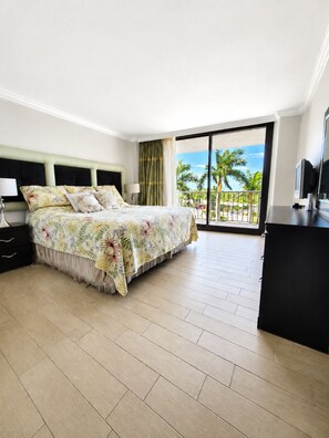 Ein Hotelzimmer mit Bett, Schreibtisch, Fernseher und einem Balkon mit Blick auf Palmen und das Meer.