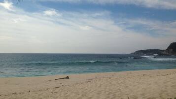 Una playa con arena, olas del océano y una costa rocosa distante bajo un cielo parcialmente nublado.