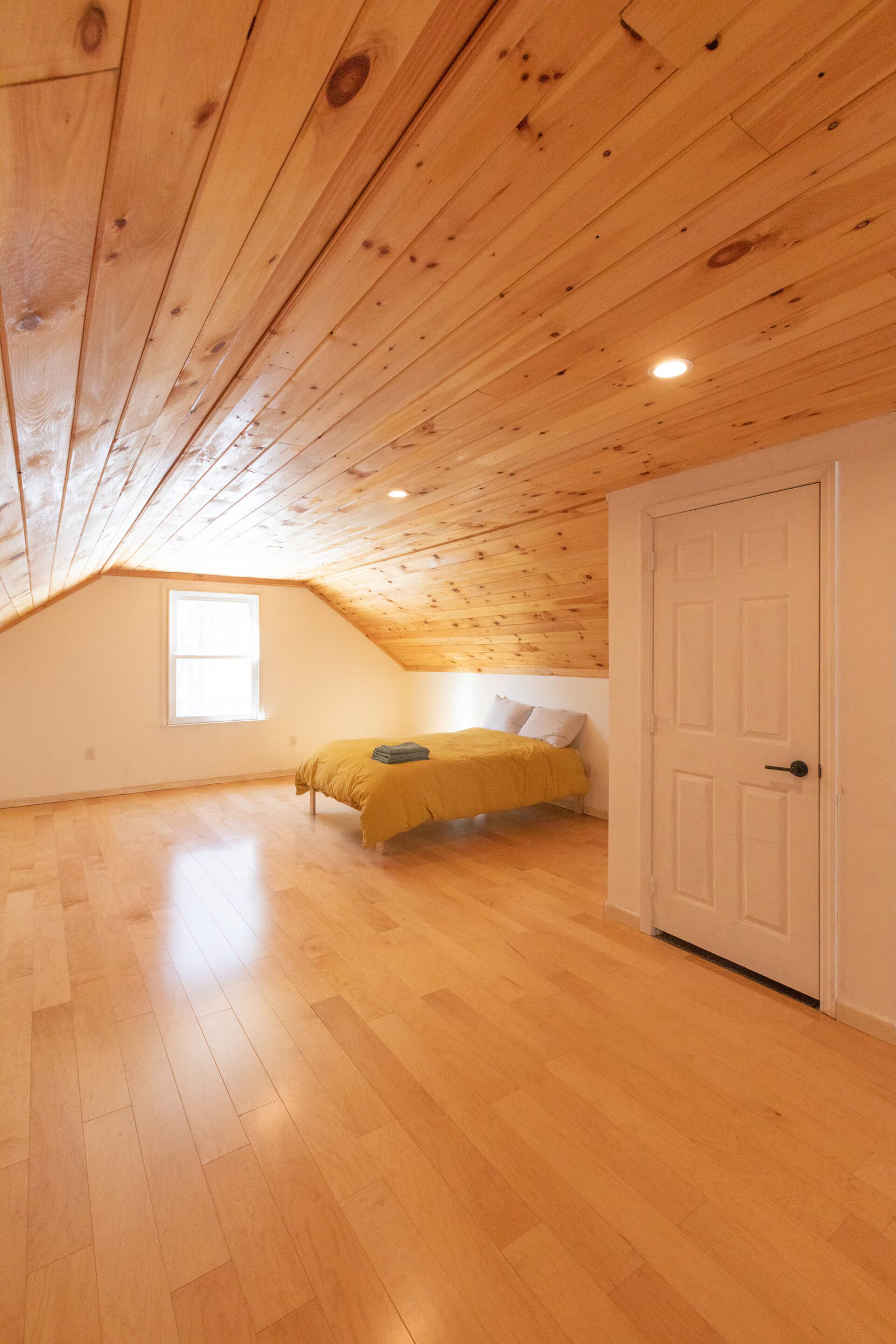A room with a sloped ceiling, wooden flooring, a bed with a yellow bedspread, a white door, and a window.