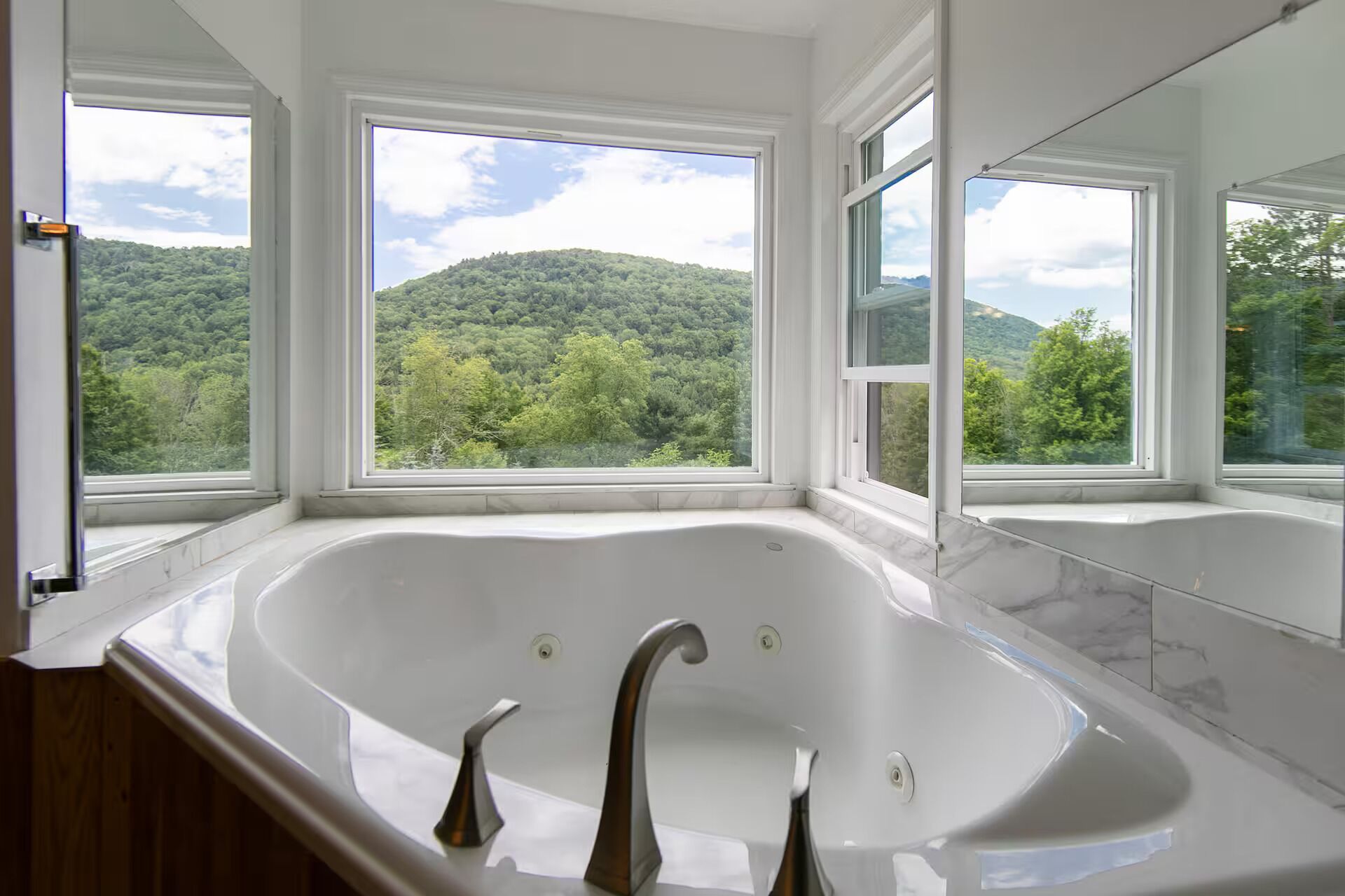A bathroom with a large bathtub, three windows, and a view of a forested hill.