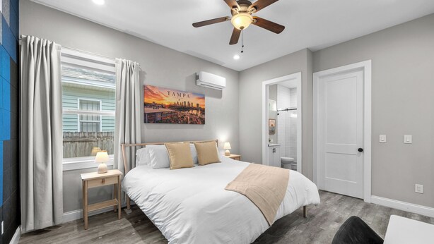 Un dormitorio con cama, ventilador de techo, ventana con cortinas, mesita de noche, lámpara, aire acondicionado instalado en la pared y puerta.