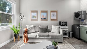 Sala de estar con un sofá gris, una mesita de centro y una planta. En la pared, hay cuadros enmarcados, una lámpara de pie y una zona de cocina con electrodomésticos.