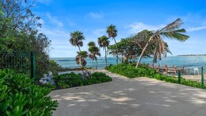 Un chemin pavé mène à une plage bordée de palmiers et délimitée par une clôture.