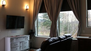 Une pièce dotée d’un téléviseur à écran plat fixé au mur, d’une commode blanche avec des tiroirs, d’un fauteuil en cuir, d’une fenêtre avec des rideaux et d’un ventilateur.