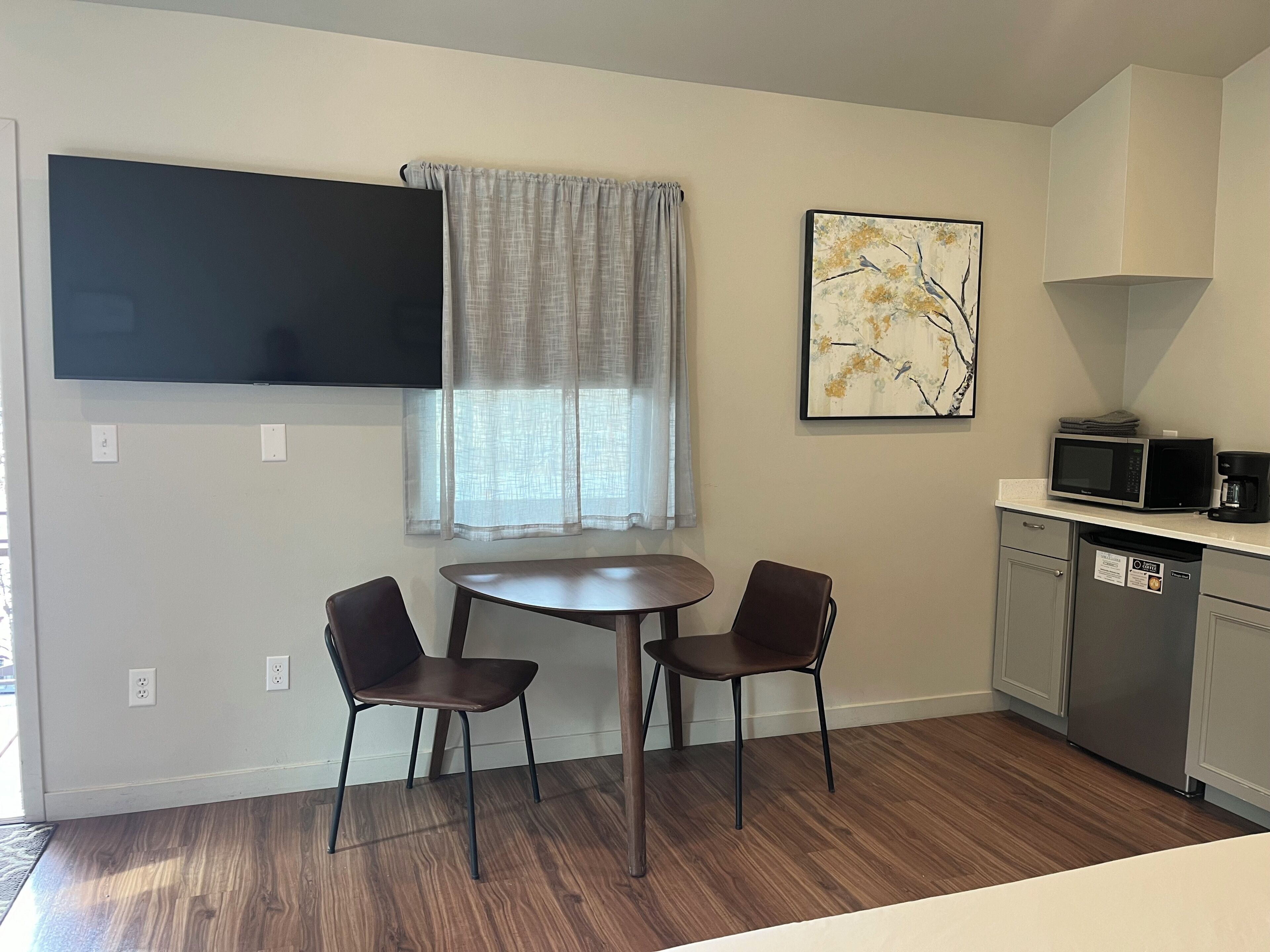 A small kitchen with a dining table and two chairs, a microwave, a coffee maker, and a painting on the wall.