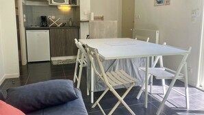 Een compacte keuken met een magnetron, koffiezetapparaat en een kleine koelkast. Op de voorgrond staat een eettafel met stoelen en een bank.