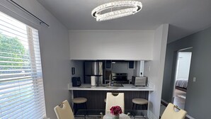 A modern kitchen with stainless steel appliances, a dining area with a glass table and white chairs, and a ceiling light fixture.