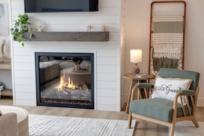 Un salon chaleureux avec une cheminée, un fauteuil confortable, une petite table d’appoint et une étagère décorative fixée au mur.