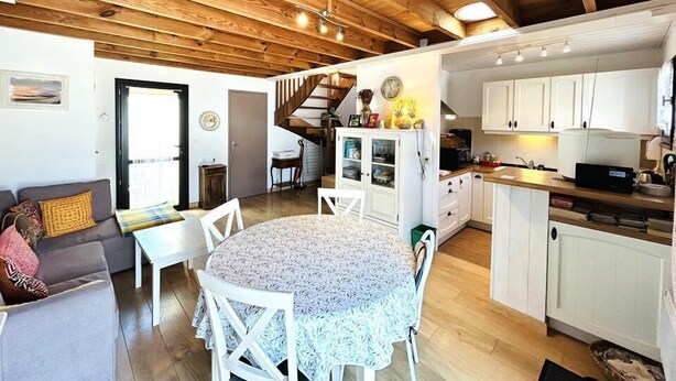 Un ambiente accogliente con soffitto in legno, zona pranzo con tavolo rotondo, cucina con mobili bianchi e una scala che conduce a un altro livello.