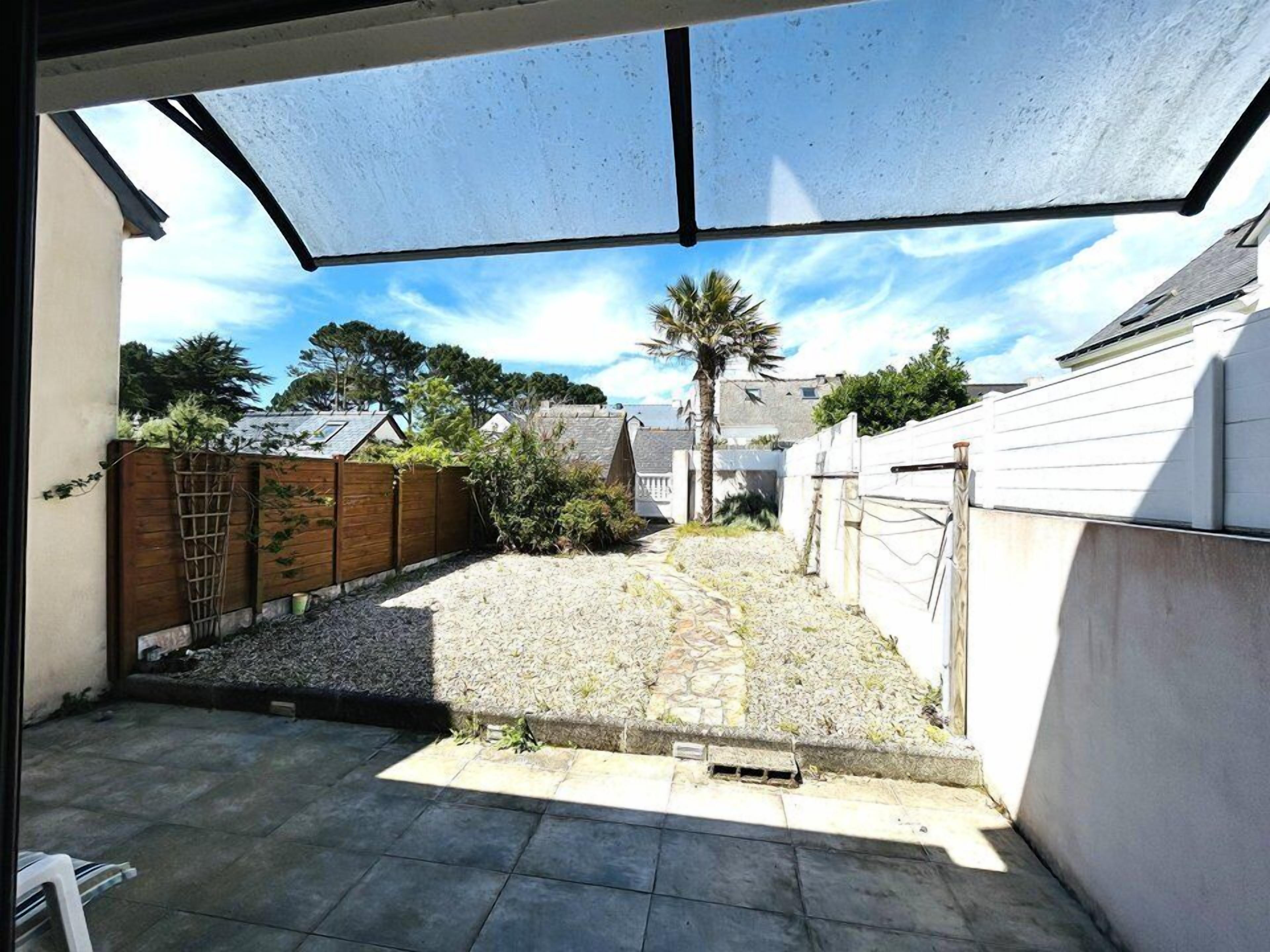 A covered outdoor area with a gravel path leading to a house, surrounded by a wooden fence and vegetation.