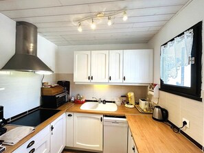 A compact kitchen with white cabinets, a stainless steel range hood, a double sink, and a window with lace curtains. - House 3 bedrooms 4 people in Guidel, only 400 m from the beach, enclosed garden, wifi (GUIDEL)