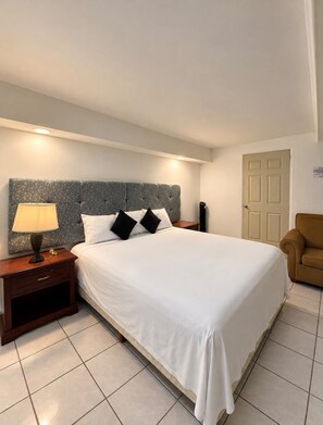A hotel room with a large bed, a nightstand with a lamp, a chair, and a door. - Hotel El Cortes (Aguascalientes)