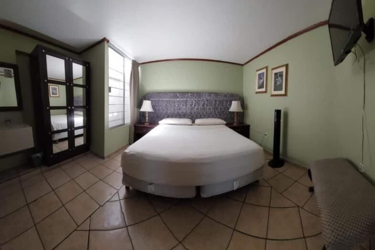 Una habitación de hotel con una cama, mesitas de noche, una silla, un televisor y una puerta que da a un baño.