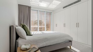 Een slaapkamer met een bed, nachtkastje, een boek, een marmeren object en een raam met zonwering.