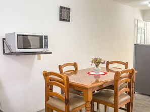 Un coin repas comprenant une table en bois, quatre chaises, un petit vase avec des fleurs, un four à micro-ondes posé sur une étagère et un réfrigérateur.