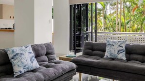 A modern living room with a grey sofa set, a coffee table with a plant, and a view of tropical foliage through large windows. - Easy Stay Pool, Esplanade Views (Cairns City)