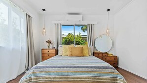 A bedroom with a large bed, wooden headboard, bedside tables, a round mirror, and a window with curtains. - The Tides - 70s Seaside Bungalow (Port Douglas)