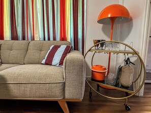 A sofa with a striped pillow, a metal cart with a lamp, and a striped curtain backdrop. - The Tides - 70s Seaside Bungalow (Port Douglas)