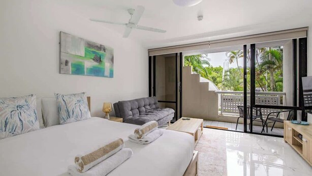 A modern hotel room with a large bed, a grey sofa, a desk, and a balcony with a view of palm trees. - Easy Stay Pool, Esplanade Views (Cairns City)