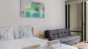 A hotel room with a bed, a grey sofa, a wooden table, and a painting on the wall. - Easy Stay Pool, Esplanade Views (Cairns City)