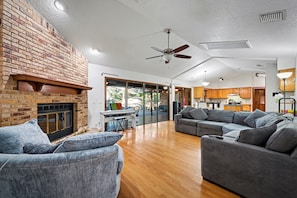 A spacious living area with a fireplace, a ceiling fan, and a kitchen in the background. - DeLand Spring House — Pool, 3/3.5, Near Blue Spring Manatees (DeLand)