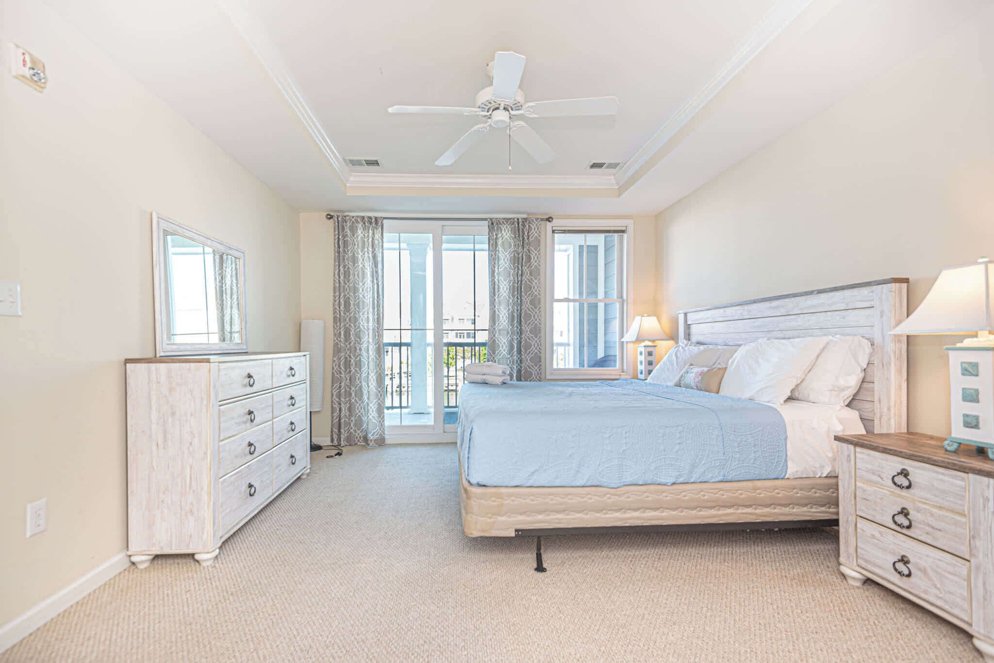 A bedroom with a bed, dresser, nightstand, ceiling fan, and large windows.