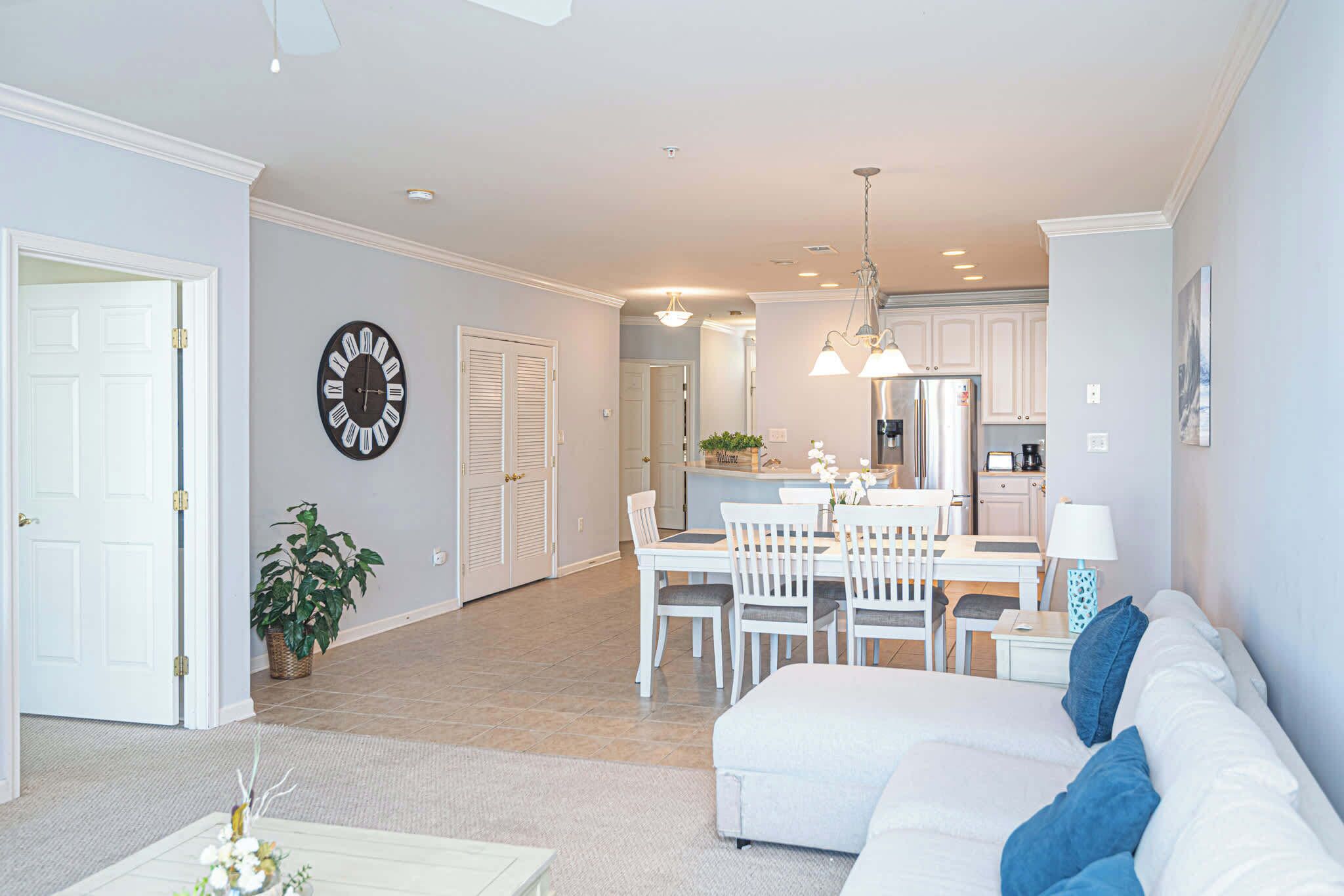 A spacious living area with a white sofa, a dining table with chairs, a kitchen in the background, and a clock on the wall.