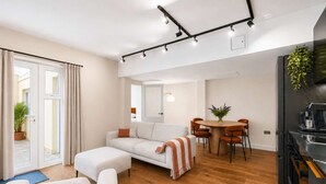 Una sala de estar moderna con un sofá, una mesa de comedor con sillas, una zona de cocina con refrigerador y un ventanal corredizo que da a un espacio exterior.