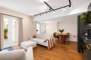 Una sala de estar moderna con un sofá, una mesa de comedor con sillas, una zona de cocina con refrigerador y un ventanal corredizo que da a un espacio exterior.