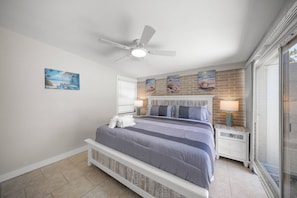 Une chambre à coucher comprenant un lit, un ventilateur de plafond, une table de chevet, une fenêtre et un mur décoré d’œuvres d’art.