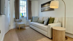 Ein modernes Wohnzimmer mit einer Couch, einem Sessel, einer Stehlampe, einem runden Holztisch und einem Fenster mit Vorhängen.