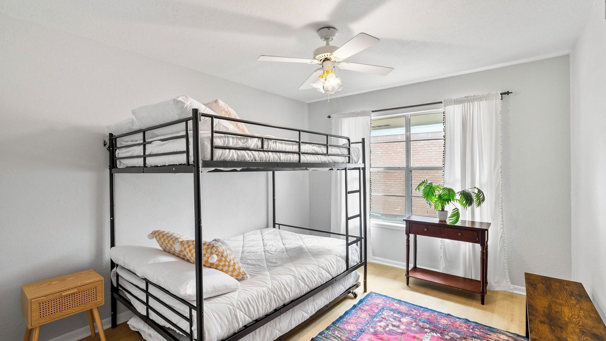Dormitorio con literas, ventilador de techo, ventana con cortinas, mesita de madera con una planta y una alfombra colorida.