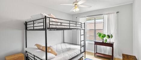 Dormitorio con literas, ventilador de techo, ventana con cortinas, mesita de madera con una planta y una alfombra colorida.
