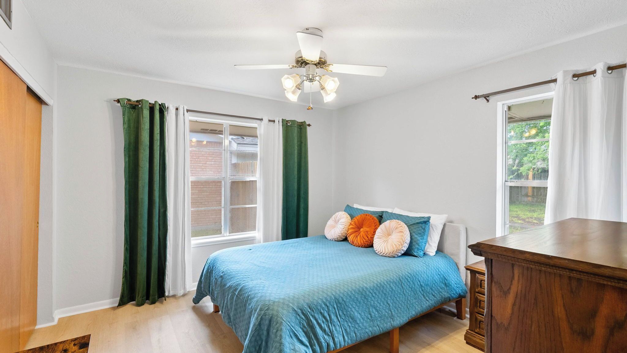 Un dormitorio con una cama, una mesita de noche de madera, un ventilador de techo y una ventana con cortinas.