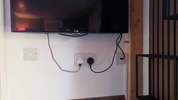 A flat-screen TV mounted on a wall with a remote control on a wooden shelf below.