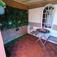 A patio with a brick floor, a round table with chairs, a potted plant, and a wall with greenery.