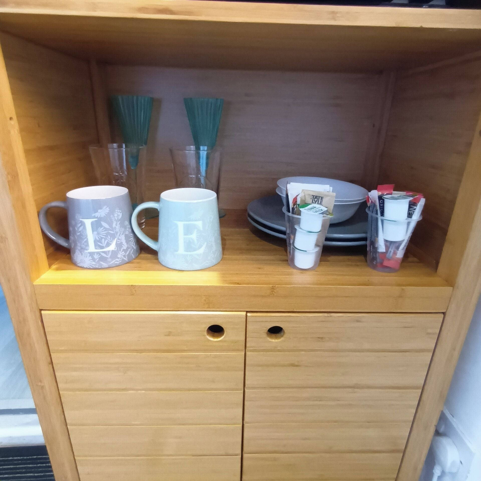 A wooden cabinet with two mugs, a glass, and a small container.