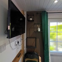 A compact room with a mounted TV, a small seating area, a window with a view, and a wall-mounted shelf with items.