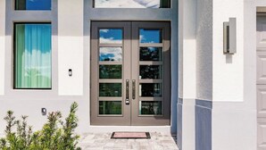 A modern building with a double door entrance, a small outdoor area with plants, and a window with a curtain. - Brisa del Mar by Avantstay Coastal Retreat + Private Pool + Lanai + BBQ Minutes to the Gulf (Naples)