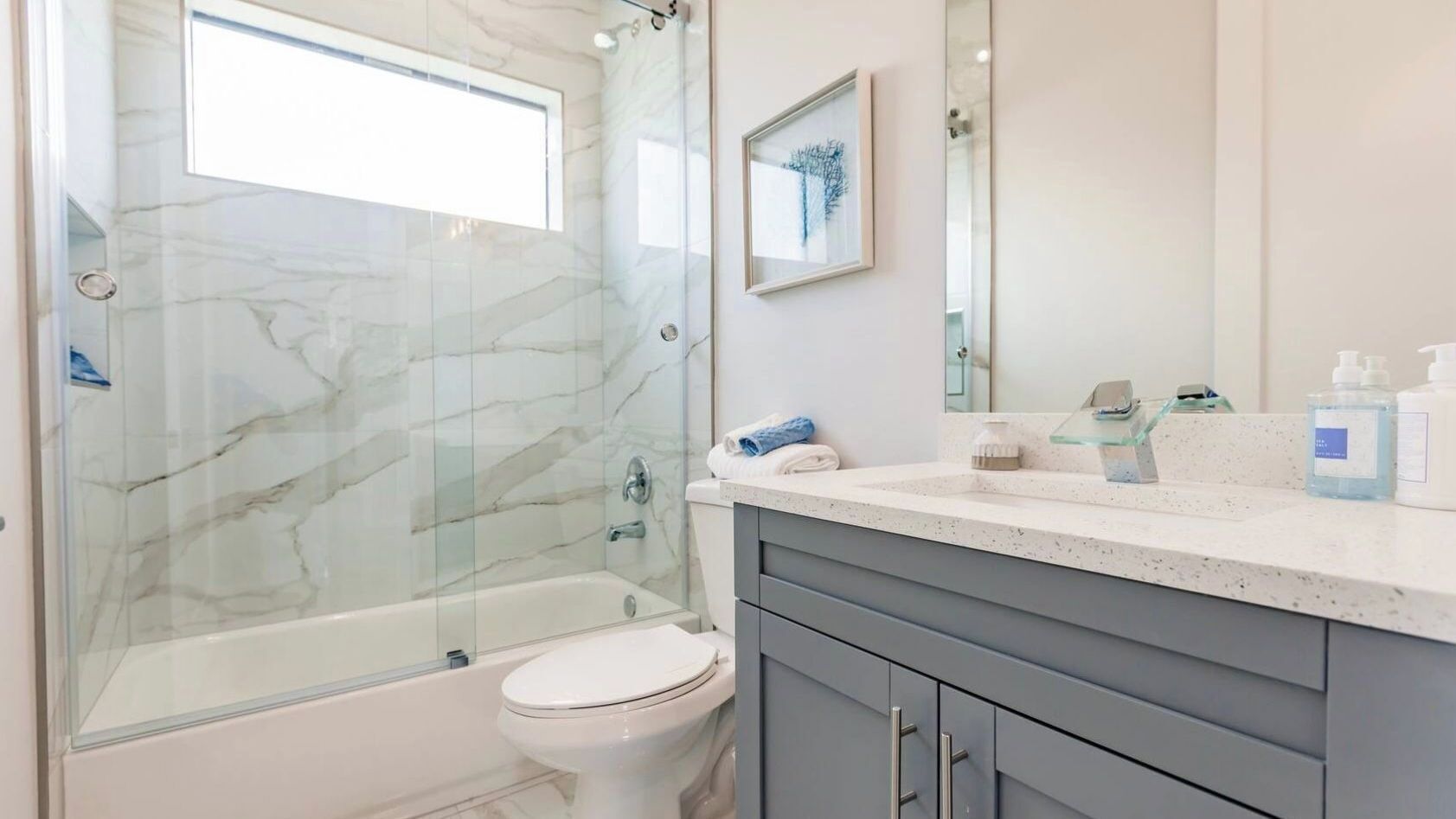 A modern bathroom with a glass-enclosed shower, a white toilet, a marble backsplash, a gray vanity, and a framed artwork on the wall.