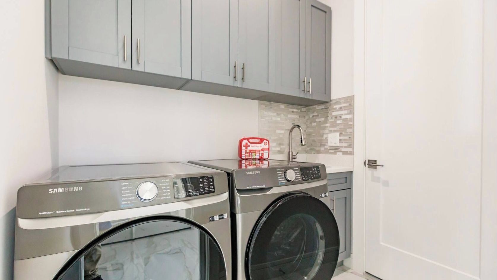 A laundry area with two Samsung washing machines, a red detergent dispenser, and grey cabinets.