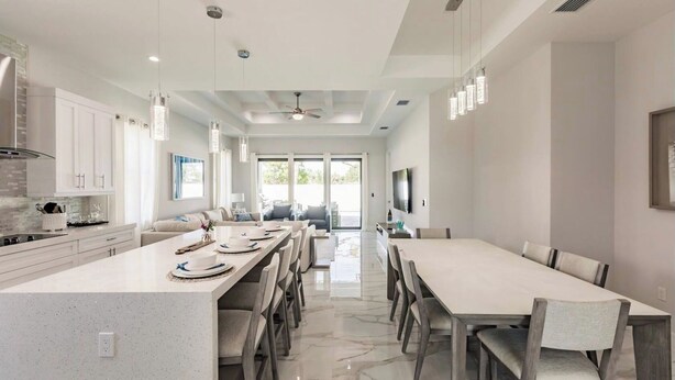 A modern kitchen with a central island, dining area with chairs, and a TV mounted on the wall. - Brisa del Mar by Avantstay Coastal Retreat + Private Pool + Lanai + BBQ Minutes to the Gulf (Naples)