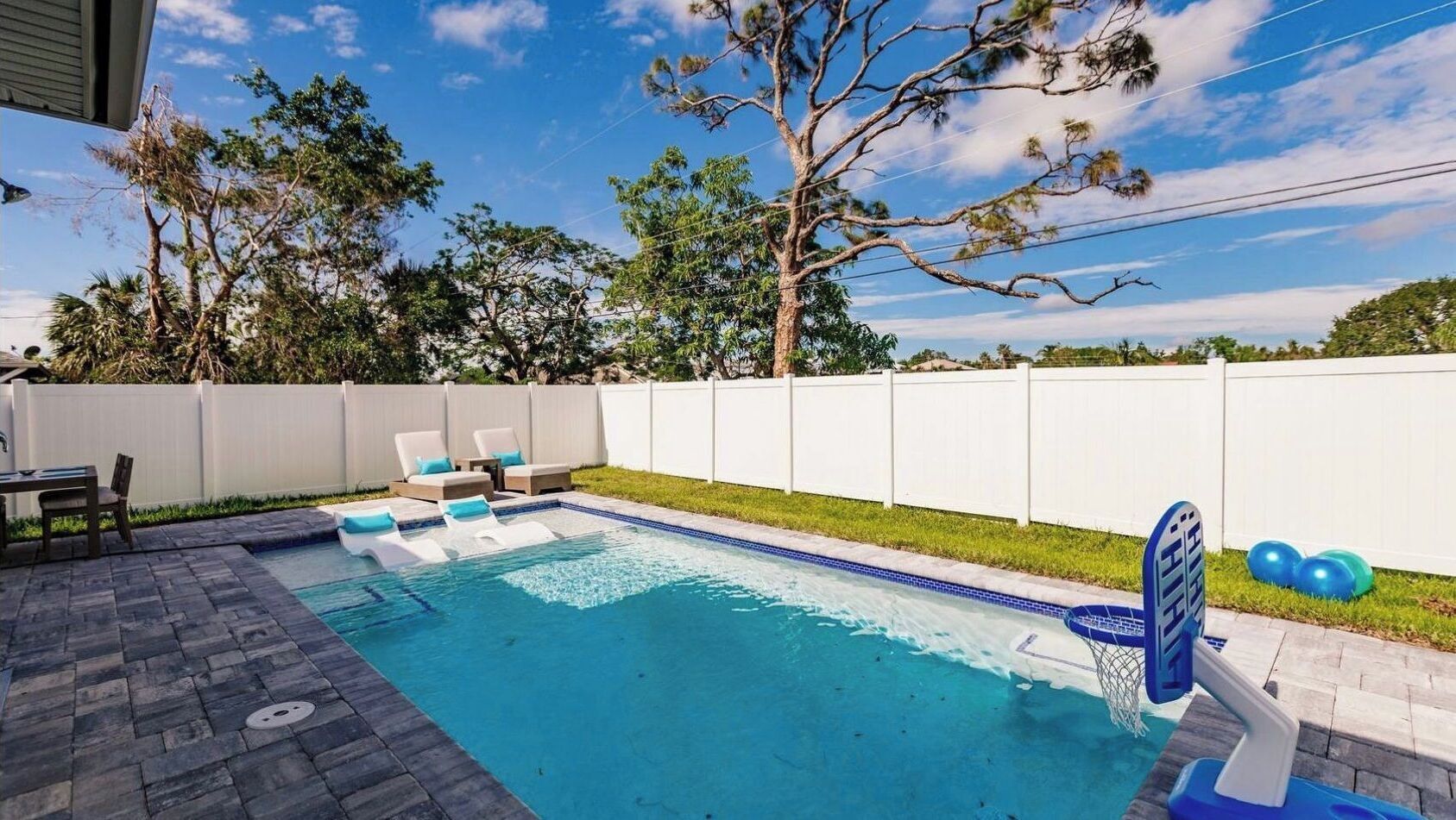 Piscina cercada por grade branca, com espreguiçadeiras e mesa.