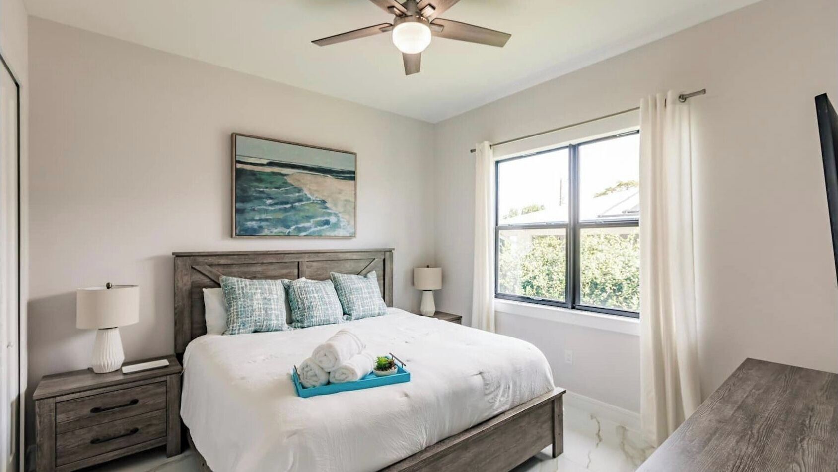A bedroom with a bed, nightstands, a ceiling fan, a window with curtains, and a painting on the wall.