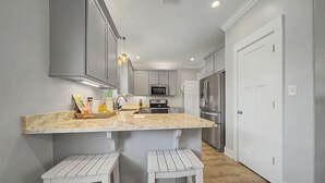 A kitchen with a marble countertop, white cabinets, a stainless steel refrigerator, and two white stools. - Welcome to Drift & Tide Cottage, your quiet coastal escape in Fort Morgan. (Gulf Shores)