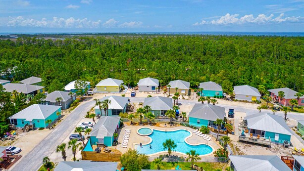 A resort with multiple blue-roofed buildings, a central swimming pool, and a surrounding forested area. - Welcome to Drift & Tide Cottage, your quiet coastal escape in Fort Morgan. (Gulf Shores)