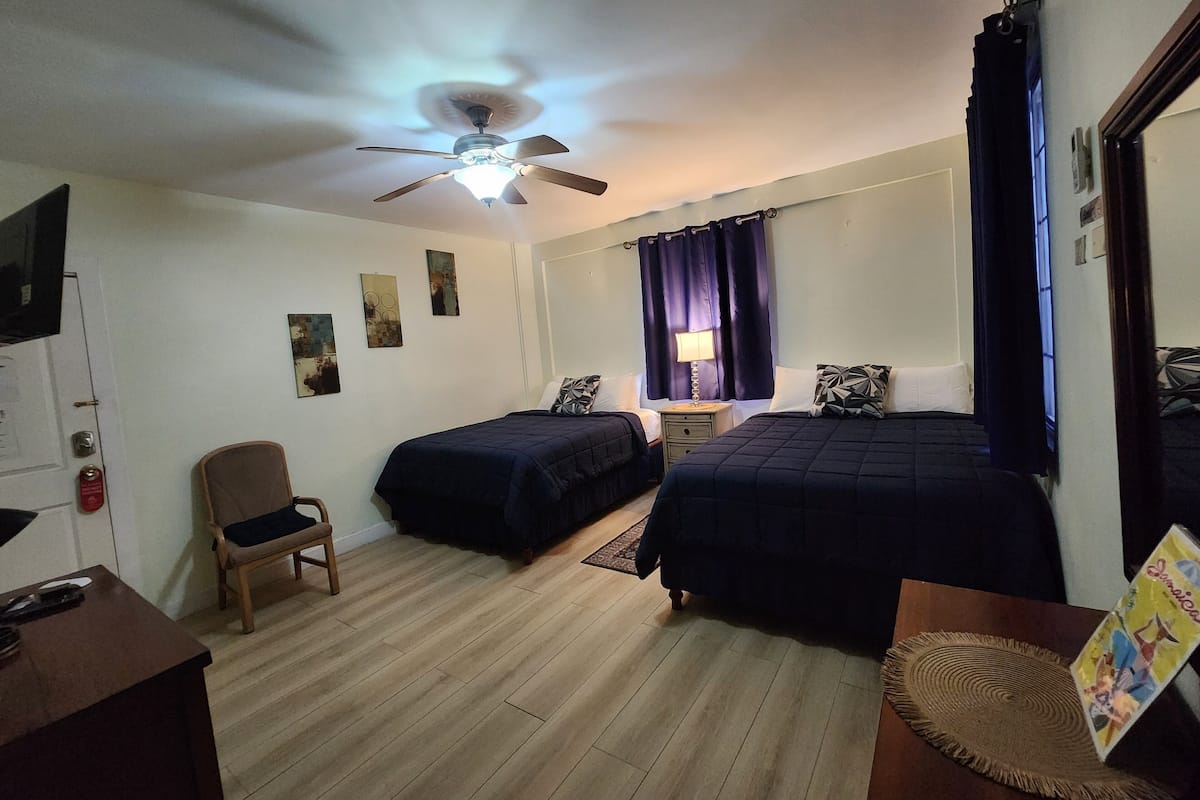 Habitación de hotel con dos camas, ventilador de techo, escritorio con silla y obras de arte en las paredes.